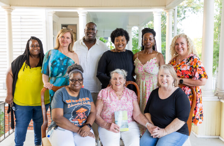 Group of diverse adults smiling and posing together on a residential porch, dressed in casual summer attire, conveying a friendly gathering.