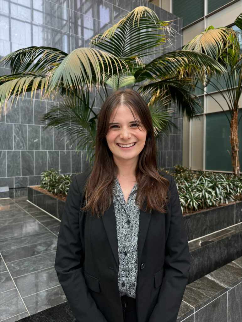 A smiling woman in a black blazer and patterned shirt stands in an indoor atrium with palm trees and marble walls.