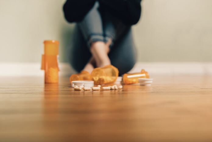 Bottles of prescription pills are spilled on a wooden floor. In the background, a person sits on the floor against a wall with arms and legs crossed.