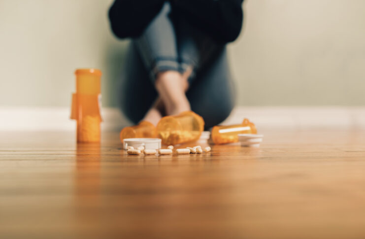 Bottles of prescription pills are spilled on a wooden floor. In the background, a person sits on the floor against a wall with arms and legs crossed.