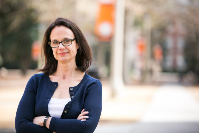 A woman with brown hair and glasses wears a white shit with a navy sweater over it. She stands with her arms crosses, with the Mercer University campus visible behind her.