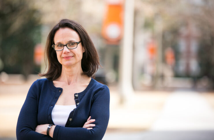 A woman with brown hair and glasses wears a white shit with a navy sweater over it. She stands with her arms crosses, with the Mercer University campus visible behind her.