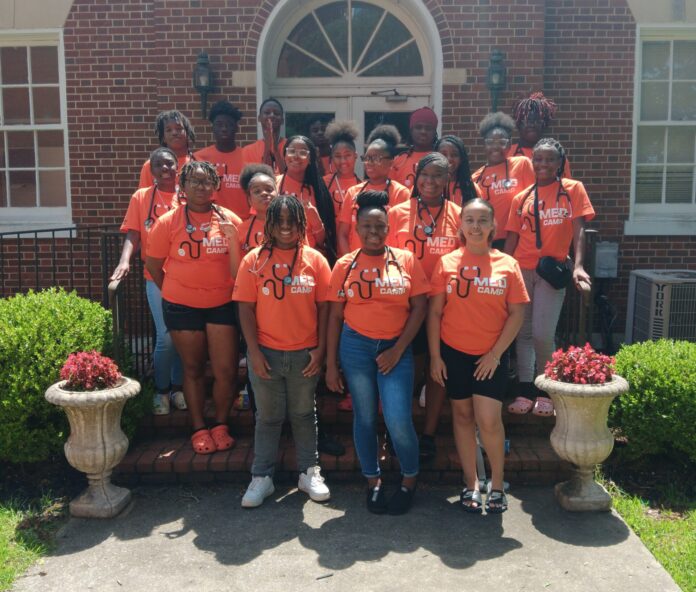 Group of students in orange T-shirts with "MED CAMP" and a stethoscope printed on them, posing in front of a brick building on a sunny day.