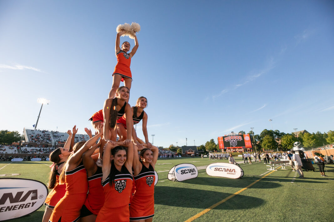 Cheerleaders wearing Mercer uniforms form a pyramid on the sidelines of a football field. A game in going on in background and people are in the stands.