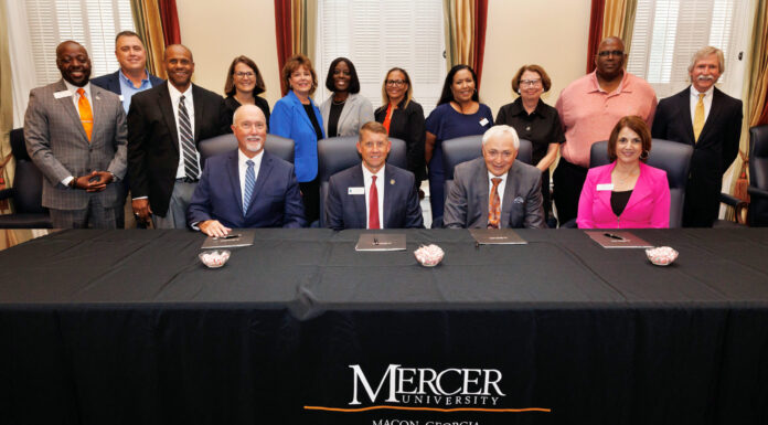 Mercer partners with Technical College System of Georgia to develop health career pathways Group photo taken during a signing ceremony at Mercer University. Three men and one woman are seated at a table covered with a black cloth that bears the university's name and location. They have documents in front of them as part of the signing process. Standing behind them are eleven other individuals, including both men and women, all dressed in business or business-casual attire, smiling for the camera.