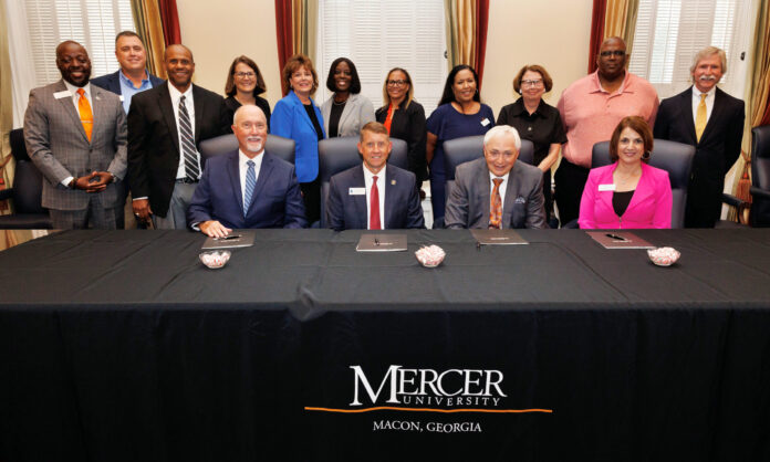 Group photo taken during a signing ceremony at Mercer University. Three men and one woman are seated at a table covered with a black cloth that bears the university's name and location. They have documents in front of them as part of the signing process. Standing behind them are eleven other individuals, including both men and women, all dressed in business or business-casual attire, smiling for the camera.