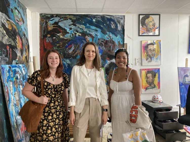 Three people standing in an art studio surrounded by colorful paintings. The individual in the center is wearing a white blouse and trousers, flanked to the left by someone in a dark floral dress and to the right by someone in a white dress. The background features vibrant abstract and figurative artworks.