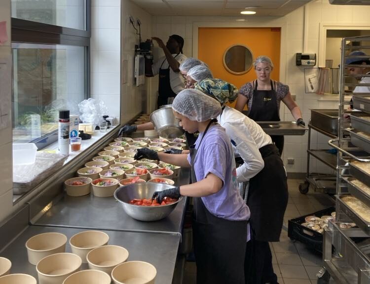 Five people in a commercial kitchen preparing food; they are wearing hair nets and aprons. Tasks include slicing vegetables and packing meals into containers. The kitchen is equipped with stainless steel countertops and various cooking utensils.