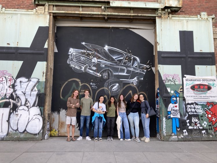 A group of seven individuals posing in front of a large mural depicting a classic car, located in an urban area with graffiti-covered walls.