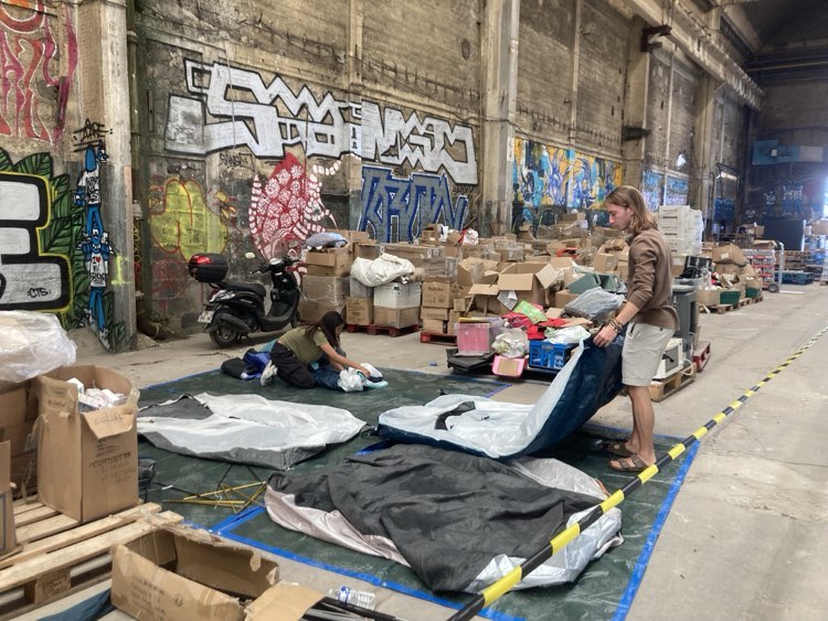 Two people are in a large industrial space with graffiti-covered walls. One person is kneeling and organizing items on the floor, surrounded by cardboard boxes, while another is standing rolling a tent. A motorbike is parked in the background. The area is sectioned off with yellow tape on the floor.