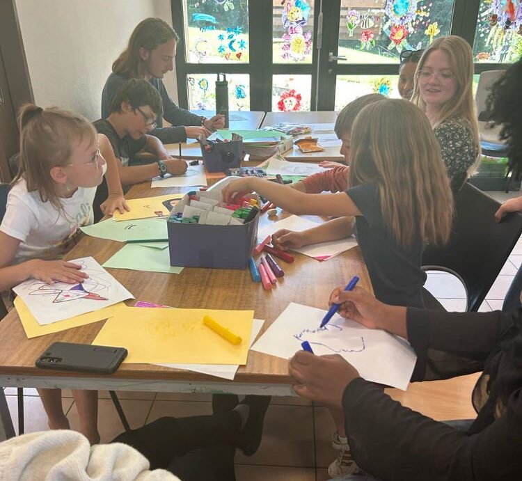 A group of children and adults seated around a table engaged in drawing and coloring activities. The table is scattered with papers, colorful markers, and a mobile phone. Large windows let natural light into the room, enhancing a bright and creative atmosphere.