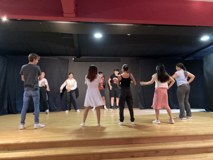 A group of people participate in a dance class, practicing steps on a wooden floor, in a room with red curtains.