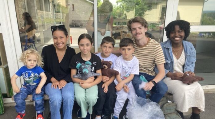 Students connect with refugees during Mercer On Mission to France Group of seven people of various ages sitting together outside a building, smiling. The group includes adults and children, some holding handmade bears, with a bag and a backpack on the ground in front of them.