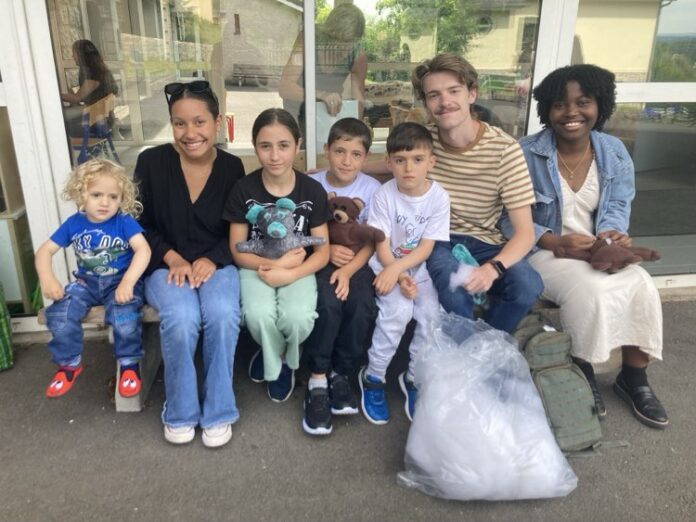 Group of seven people of various ages sitting together outside a building, smiling. The group includes adults and children, some holding handmade bears, with a bag and a backpack on the ground in front of them.