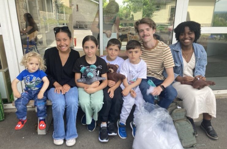 Group of seven people of various ages sitting together outside a building, smiling. The group includes adults and children, some holding handmade bears, with a bag and a backpack on the ground in front of them.