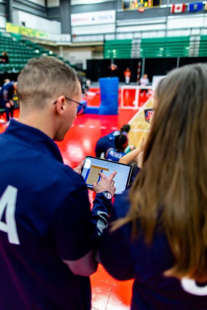 A man and woman stand together and look at a laptop, held by the man. Behind them, an indoor sports facility can be seen.