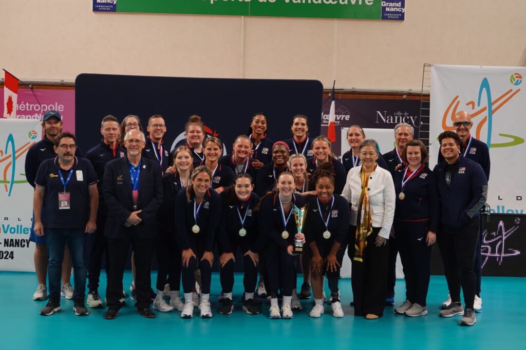 A large group of men and women pose for a photo inside an indoor sports facility, with most of them wearing athletic track suits and medals around their necks.