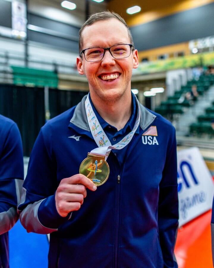 A man wears a navy blue USA track suit and holds up a gold medal on his neck.