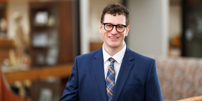 Professional headshot of a man in a navy blue suit and plaid tie, wearing black-framed glasses. He has short curly brown hair and is smiling, standing indoors with a blurred background of office furniture and warm lighting.