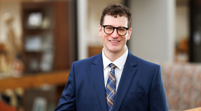 School of Medicine names new director of Master of Family Therapy program Professional headshot of a man in a navy blue suit and plaid tie, wearing black-framed glasses. He has short curly brown hair and is smiling, standing indoors with a blurred background of office furniture and warm lighting.