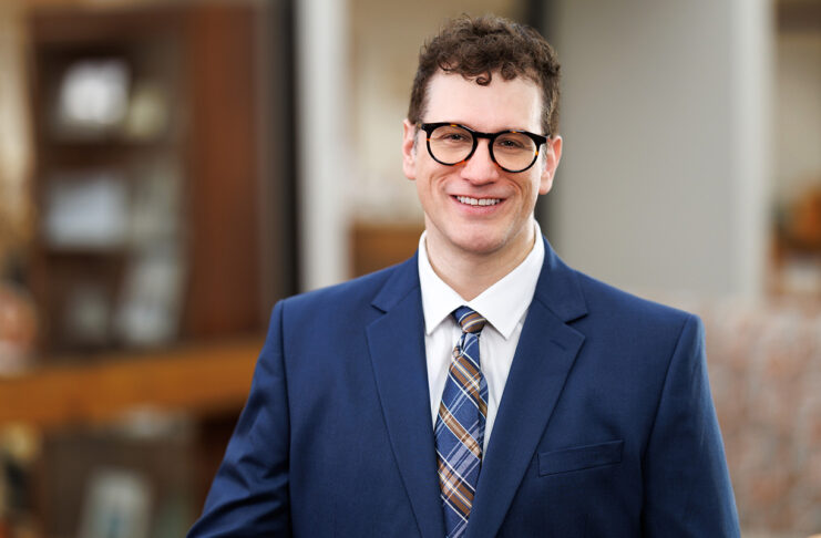 Professional headshot of a man in a navy blue suit and plaid tie, wearing black-framed glasses. He has short curly brown hair and is smiling, standing indoors with a blurred background of office furniture and warm lighting.