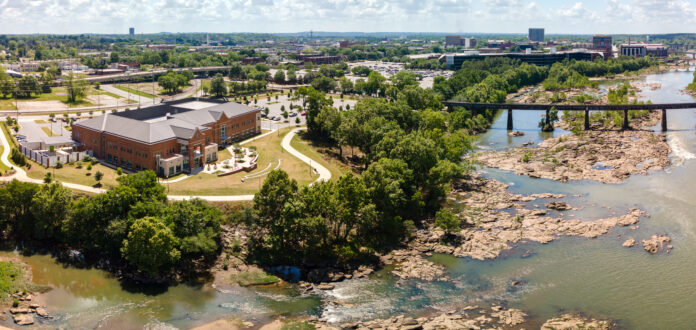 An aerial view of a scenic area featuring a large building, a river with rocky terrain, a bridge crossing the water, and surrounding greenery. enhancing the natural beauty.