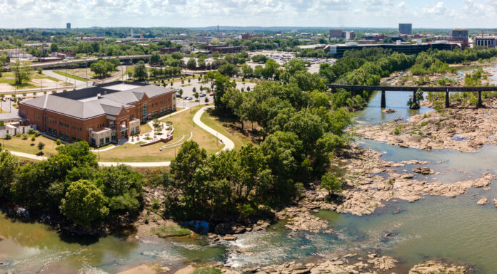 Faculty and Staff Notables | September 2024 An aerial view of a scenic area featuring a large building, a river with rocky terrain, a bridge crossing the water, and surrounding greenery. enhancing the natural beauty.