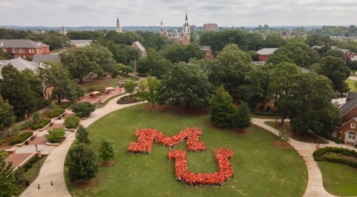 Mercer announces record fall enrollment An aerial view of a university campus with a large group of people forming the shape of the letters "MU" on a lawn. The campus is surrounded by lush green trees and several brick buildings with pointed roofs. The sky is overcast.