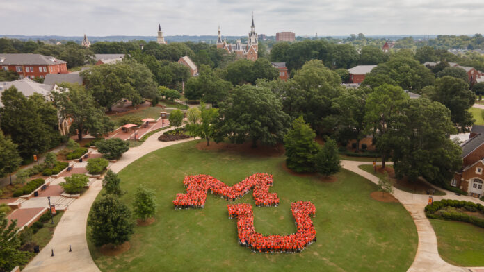 An aerial view of a university campus with a large group of people forming the shape of the letters "MU" on a lawn. The campus is surrounded by lush green trees and several brick buildings with pointed roofs. The sky is overcast.