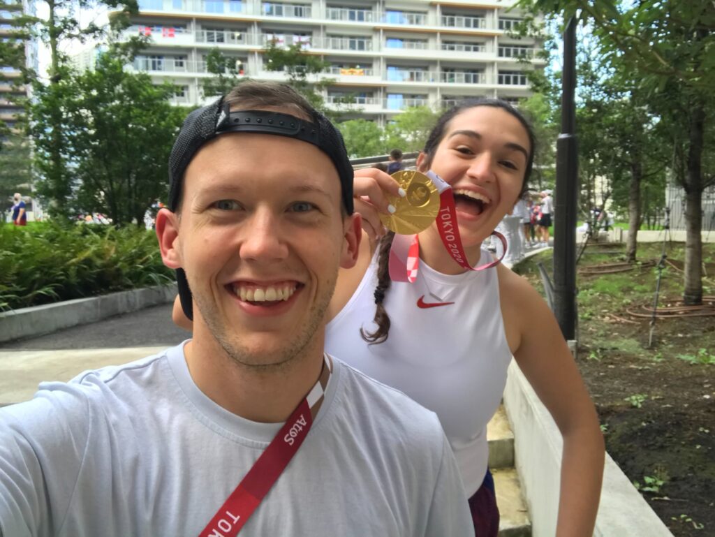 A man takes a selfie of himself, wearing a white shirt and backward baseball cap, and a women behind him in a white shirt holding a gold medal that's around her neck. Behind them are trees and a tall building.