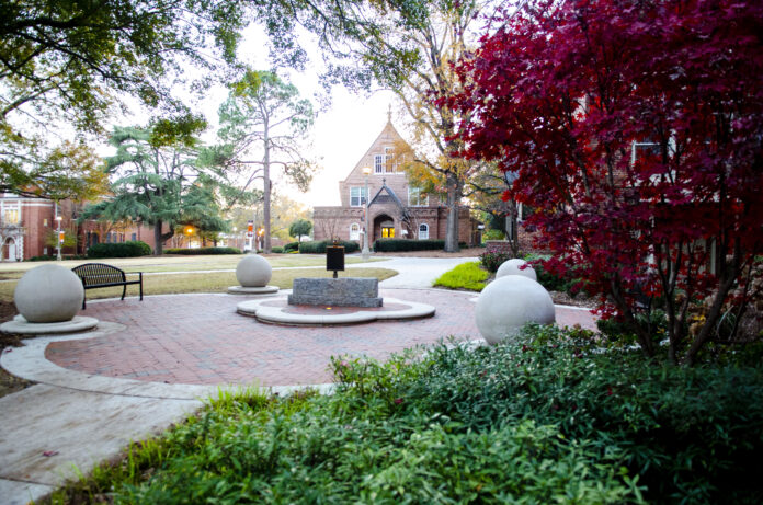 Peaceful courtyard surrounded by greenery and red brick buildings. The scene includes a circular paved area with large spherical stone sculptures and a central plaque or marker. The buildings in the background have an old, academic appearance. The trees surrounding the area have both evergreen and autumn-colored leaves.