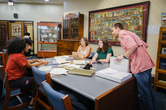 Four individuals engaged in a discussion around a table at a library, surrounded by books and artwork on the walls. They are using laptops, books, and notepads.