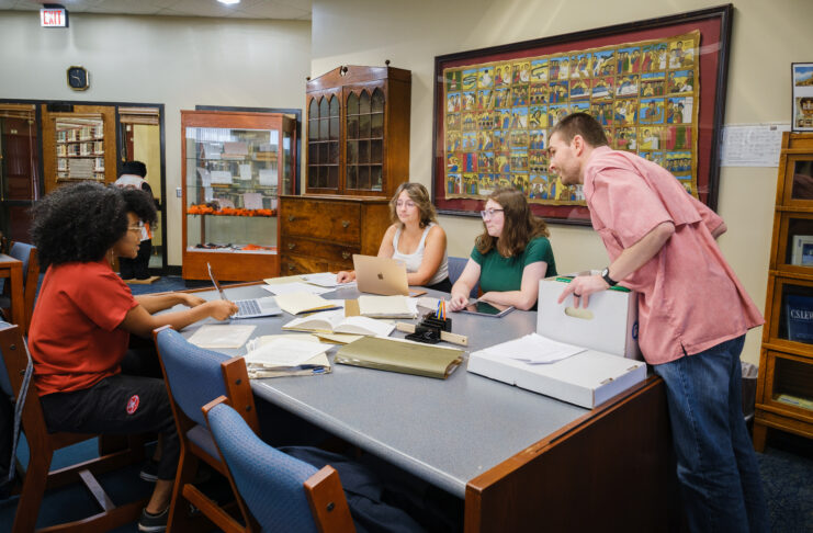 Four individuals engaged in a discussion around a table at a library, surrounded by books and artwork on the walls. They are using laptops, books, and notepads.