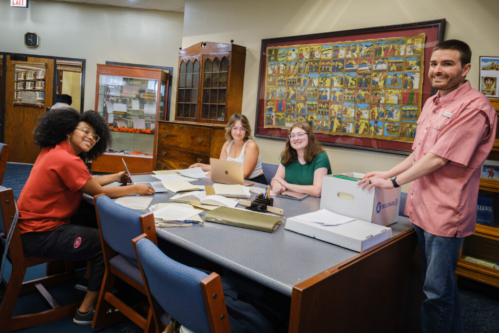 Four individuals look at materials in a library room, with books and papers on the table. One person stands while three others are seated around the table.