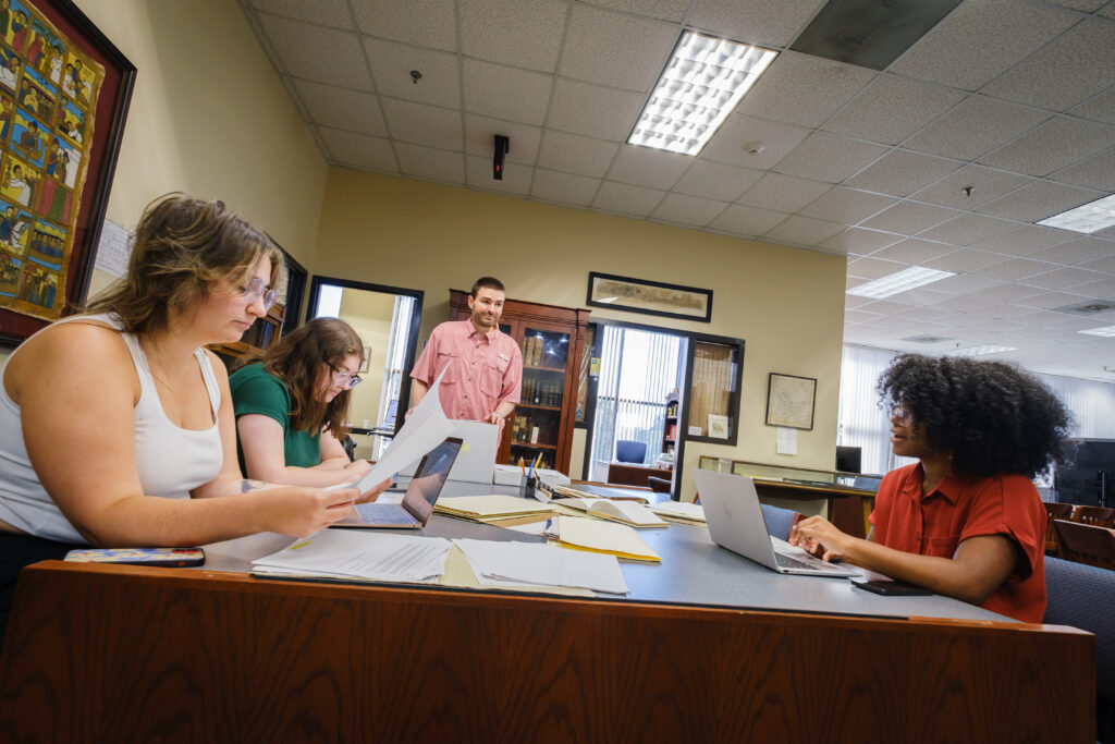 Four individuals engaged in a discussion around a table at a library, surrounded by books and artwork on the walls. They are using laptops, books, and notepads.