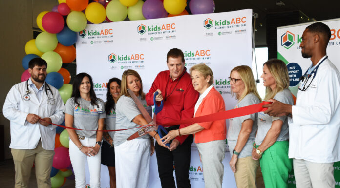 KidsABC celebrates Mountain Lakes Medical Center’s Emergency Department as part of the Pediatric Emergency Care Project A group of people is gathered for a ribbon-cutting ceremony at the Mountain Lakes Medical Center. The backdrop features the "kidsABC" logo. The individuals are dressed in a mix of professional and casual attire, with some wearing medical coats. They are smiling as they cut a red ribbon, with colorful balloons decorating the background.