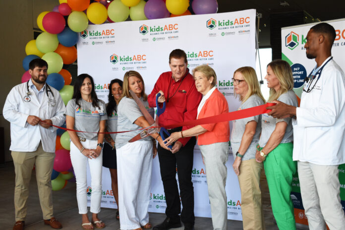 A group of people is gathered for a ribbon-cutting ceremony at the Mountain Lakes Medical Center. The backdrop features the "kidsABC" logo. The individuals are dressed in a mix of professional and casual attire, with some wearing medical coats. They are smiling as they cut a red ribbon, with colorful balloons decorating the background.