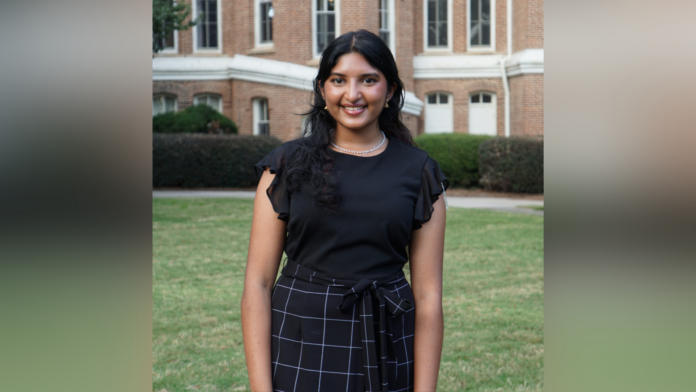 A woman smiles for a photo while standing in front of a brick academic building and lush green grass. She wears a black blouse and black checkered skirt.