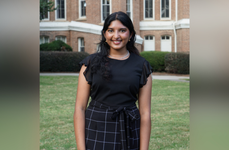 A woman smiles for a photo while standing in front of a brick academic building and lush green grass. She wears a black blouse and black checkered skirt.