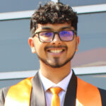 Headshot of Adil Ahmed wearing a black suit, white button up shirt and orange tie. He has an orange stole around his neck and is wearing glasses.