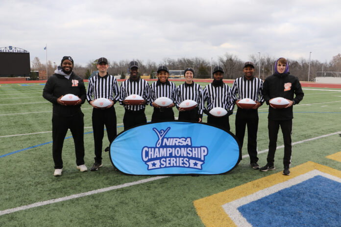 Six people wearing white and black striped official uniforms stand on a football field in front of a sign that says, "NIRSA Championship Series." They are flanked by two people wearing all black.