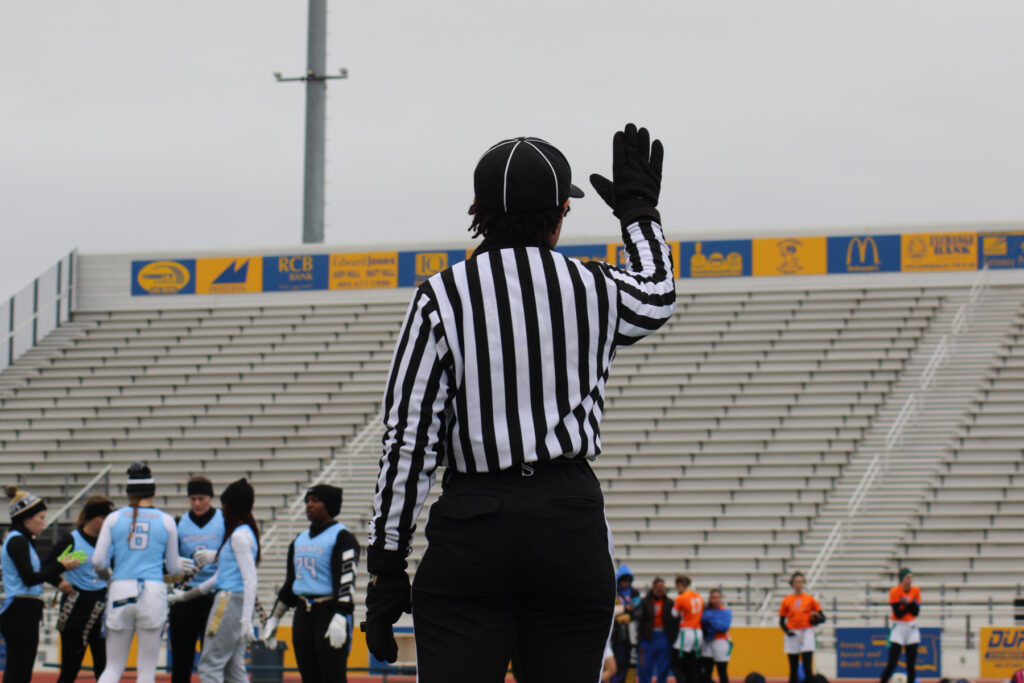 The back of an official lifting her hand during a flag football game. Players are seen on the field.