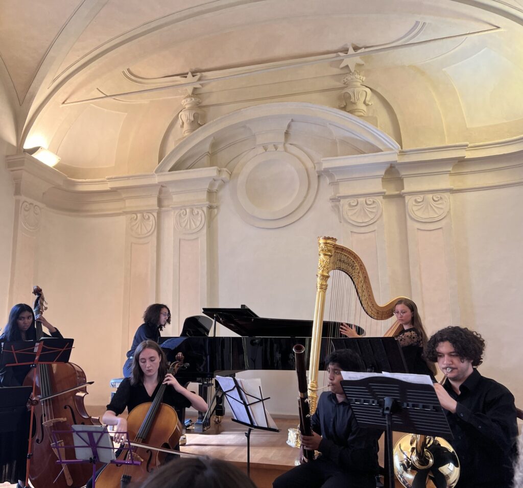 Musicians performing in an ornate room with a harp, piano, and strings, under a vaulted ceiling.