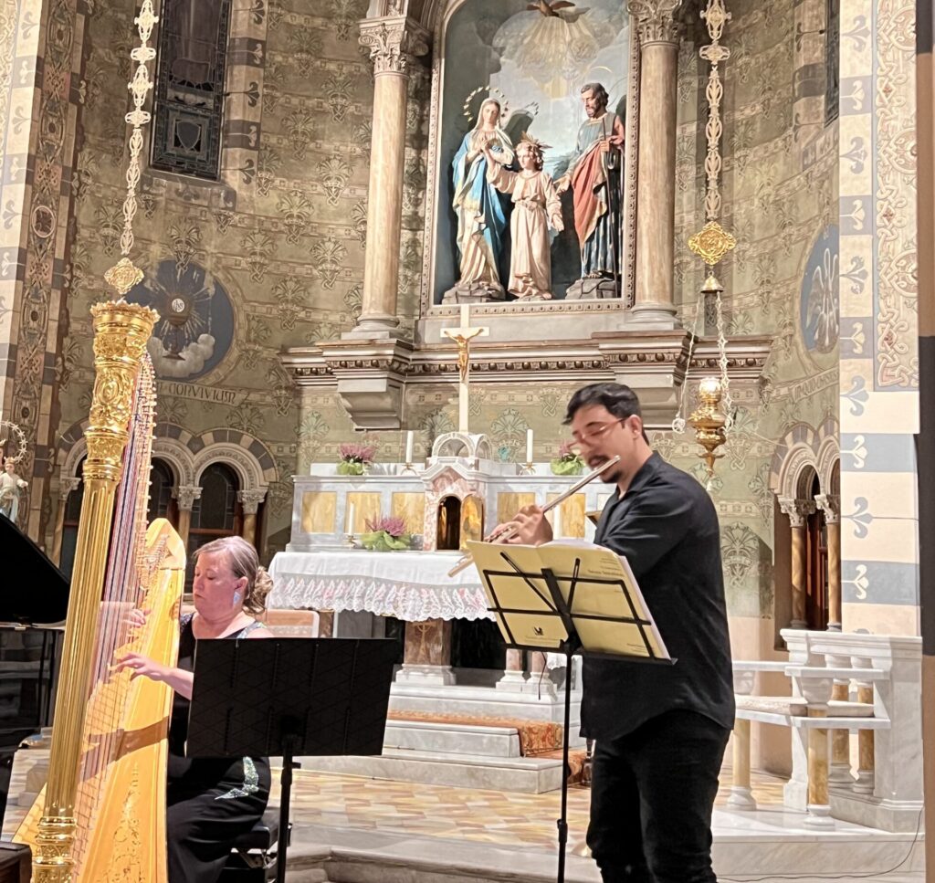 A man stands to play flute with a harpist seated beside him, both performing inside a richly decorated church with frescoed walls and an ornate altar.