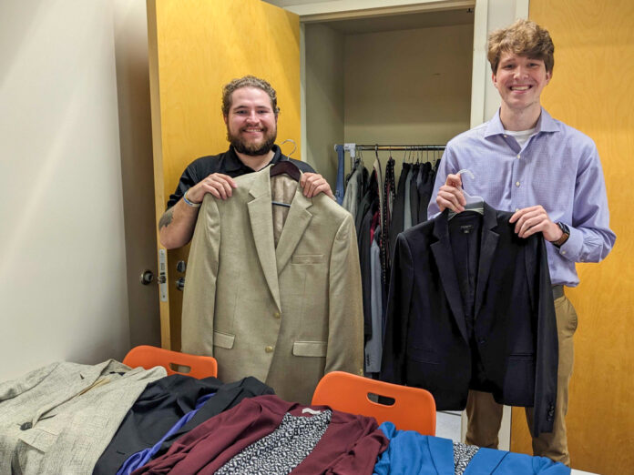 Two Mercer students smile while holding up blazers. More blazers are laid out on a desk in front of them, and others are hung in a closet behind them.