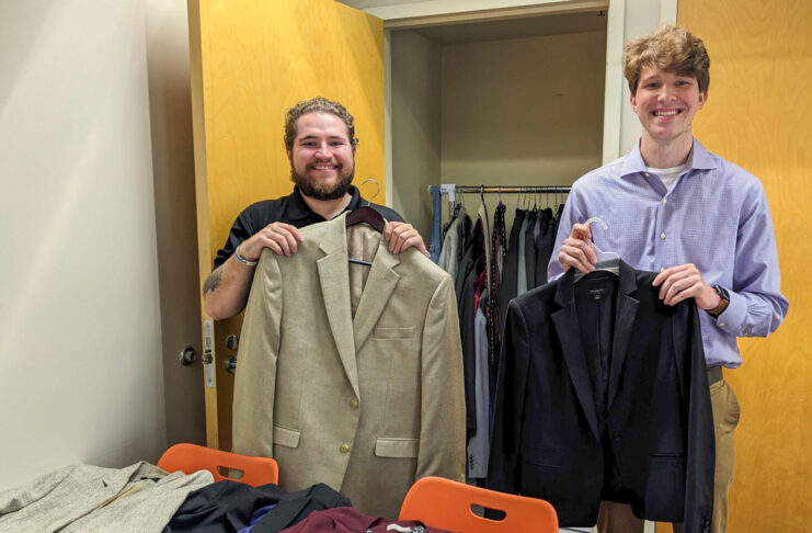 Two Mercer students smile while holding up blazers. More blazers are laid out on a desk in front of them, and others are hung in a closet behind them.