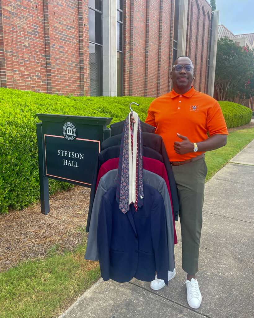 Nnamdi Onyekwuluje wears an orange Mercer polo shirt while holding a hanger with multiple blazers and ties. He stands in front of a building and a sign that says, "Stetson Hall."