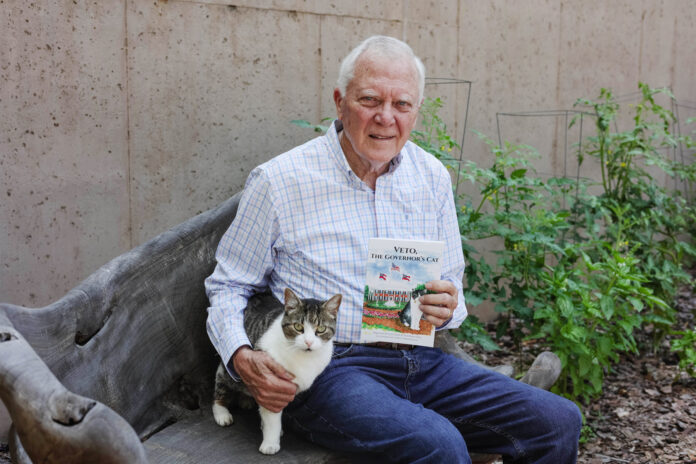 Nathan Deal sits on a bench outside holding a book titled, "Veto, the Governor's Cat." A white and brown cat is seated next to him. Tomato plants are in the background.