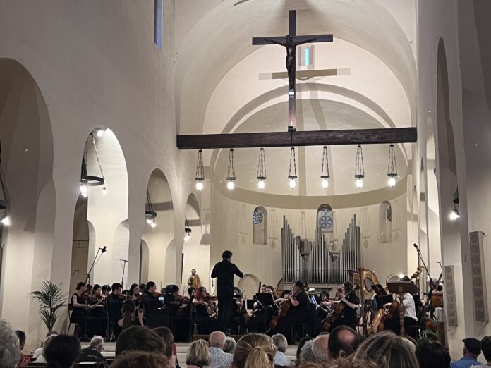 An orchestra performs in a church with a large cross and organ pipes in the background, under the observation of an attentive audience.