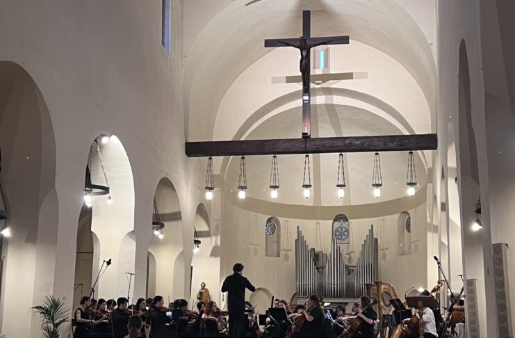 An orchestra performs in a church with a large cross and organ pipes in the background, under the observation of an attentive audience.